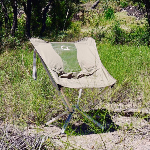 An olive green folding camp chair with a clear back panel sits in a grassy outdoor wooded area. Limited Edition MystiQ Olive Green- CLIQ