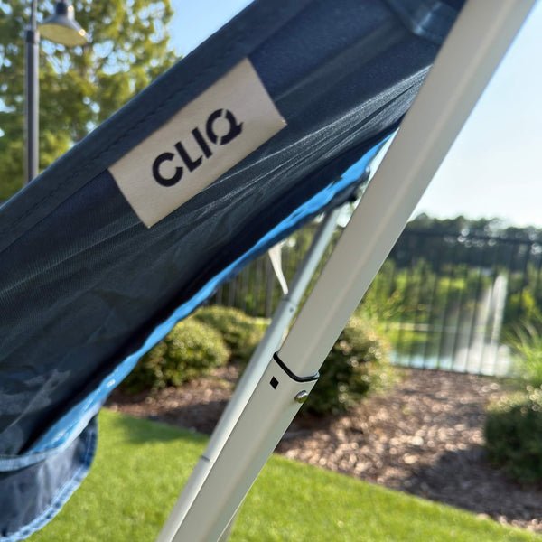 Close-up of a dark blue mesh canopy with a white metal frame and a visible CLIQ brand tag, set outdoors on grass with trees and a fence in the background. Limited Edition MystiQ Blue - CLIQ