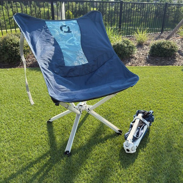 A blue folding camp chair with a white metal frame is set on green grass outdoors, with its folded version placed beside it. A black fence and landscaped garden beds are visible in the background. Limited Edition MystiQ Blue - CLIQ