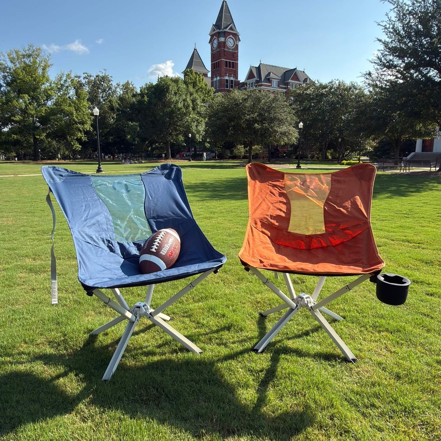 Two folding camp chairs on a grassy lawn with trees and a clock tower building in the background. The left chair is blue with a football resting on it, and the right chair is orange with a cup holder on the side. - Limited Edition MystiQ Chairs - CLIQ
